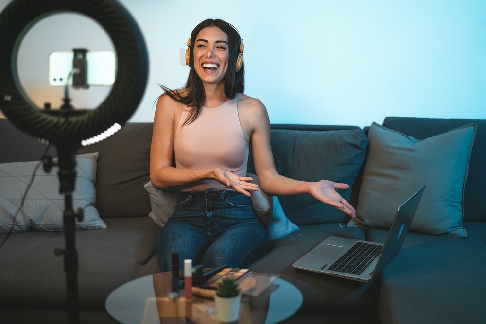 Brunette young woman sitting on a brown couch with headphones and a laptop open. In front of her is a table with makeup and a ringlight with her phone recording.