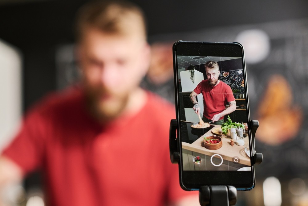 White man in a red shirt cooking on a live stream while the phone records it.