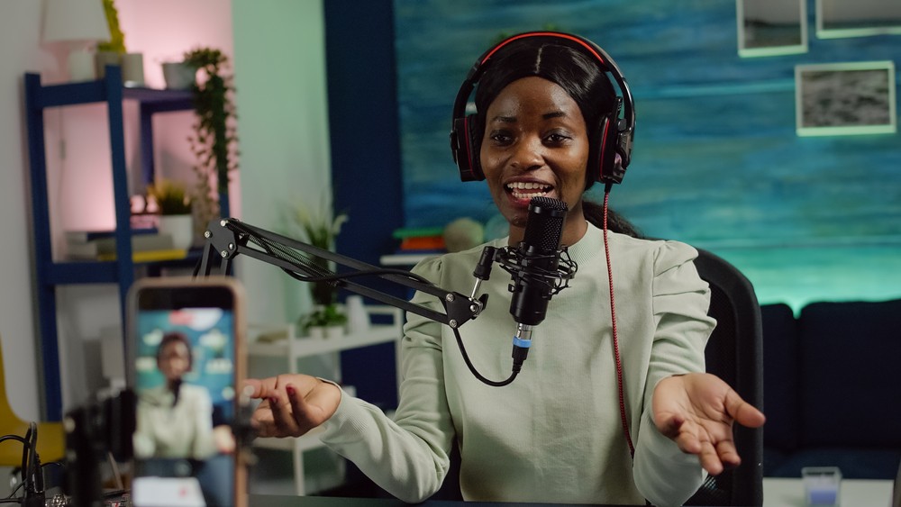 Young Black woman sitting in front of a microphone with headphones on recording herself on a livestream.