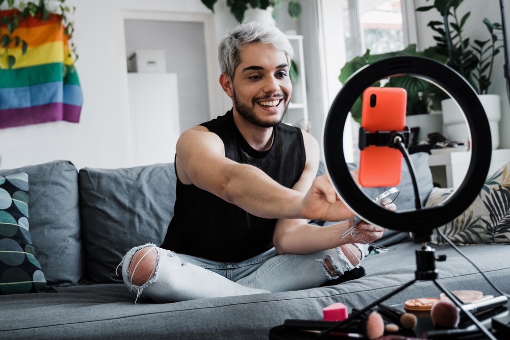 Young gay man with platinum hair sitting in front of a tripod applying makeup.