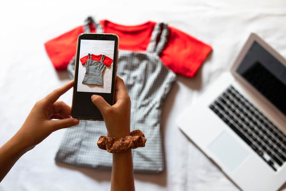 Woman's hands taking a picture of a checkered top. Her laptop is sitting on the bed next to the top.