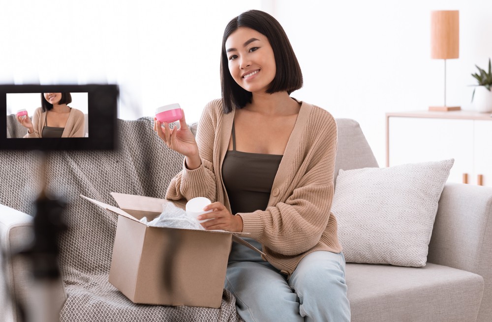Young asian woman olding a pink skin care container and smiling at the camera on her tripod.