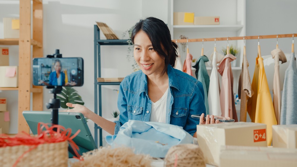 Asian woman with packing materials on her desk and a rack of clothing behind her talking on a livestream.