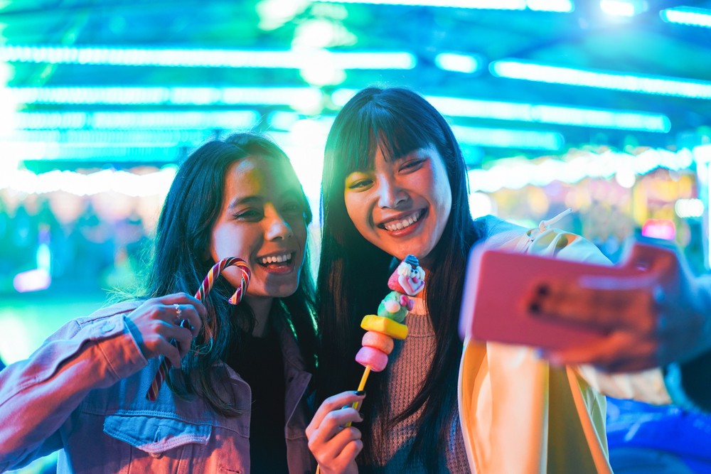 Two young women taking a selfie while holiding up candy treats in front of colorful lights in the background.