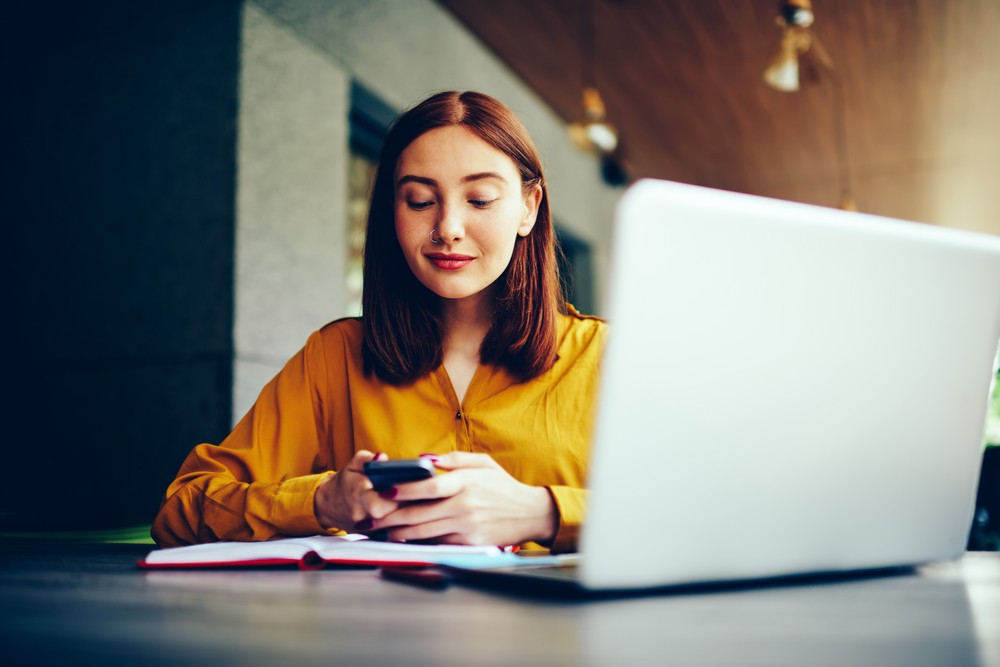 Young woman with short red hair sitting at a table with her laptop and a notebook open checking her phone.