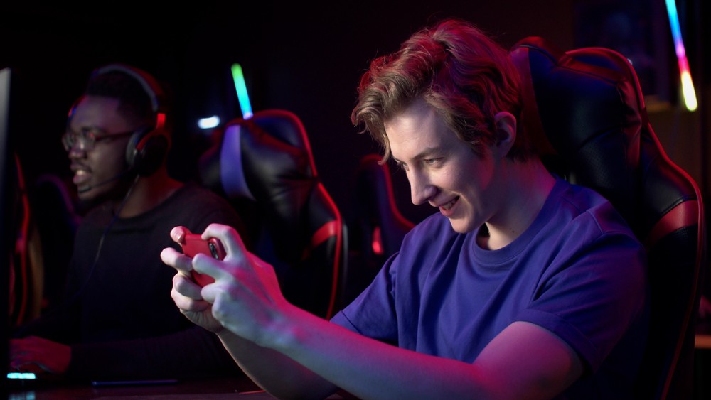 Young blonde man recording himself playing in a gaming chair. There is a black young man sitting next to him gaming.