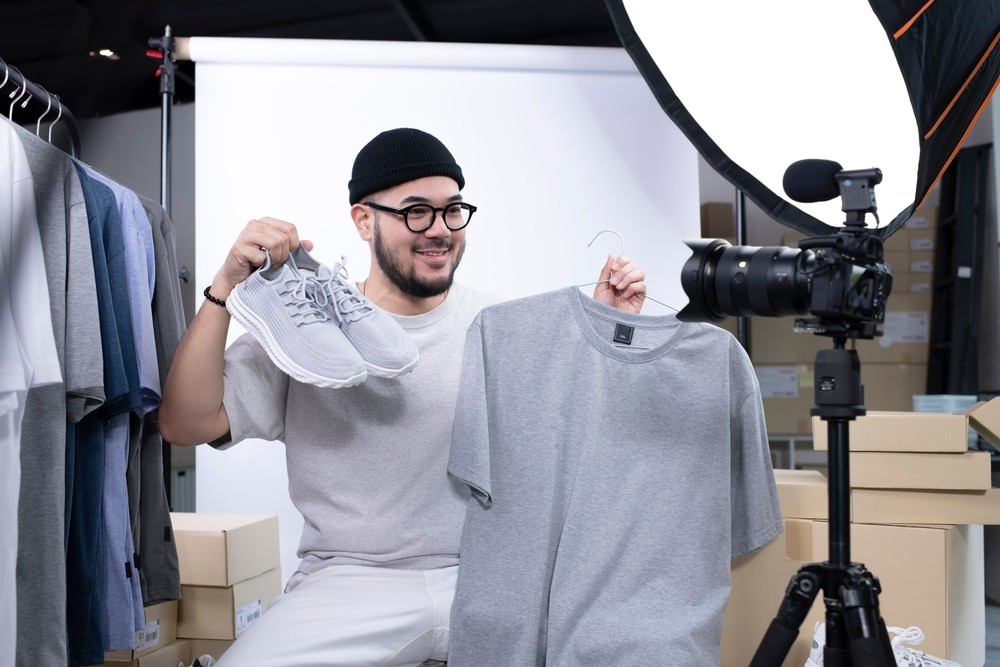 Young man holding up shoes and shirt in front of a camera with a backdrop and clothes on a rack in the background.
