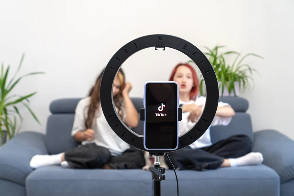 Two young women sitting on a grey couch in front of a tripod making a TikTok video.