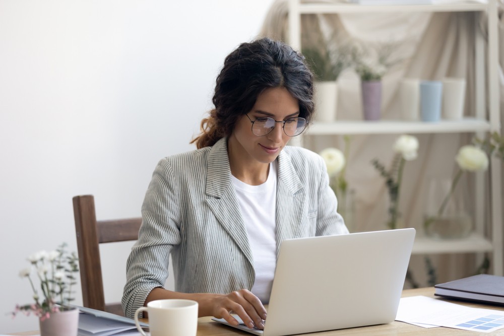 Brunette woman wearing a blazer typing on her laptop in a home office with flowers on the desk.
