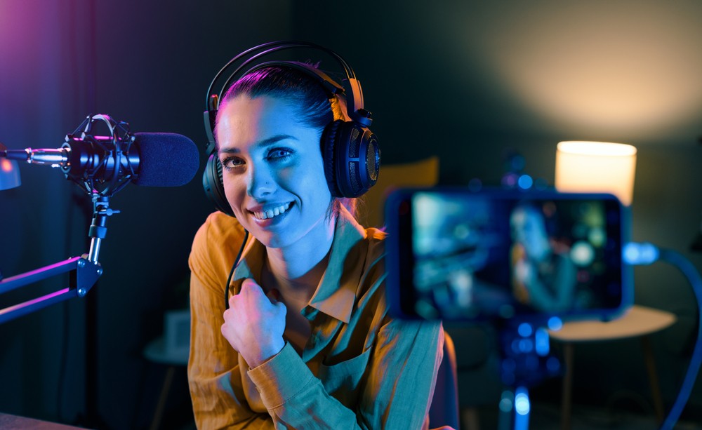 White woman sitting in front of her phone with heaphones and a microphone while recording. Purple and blue lights are in the background.