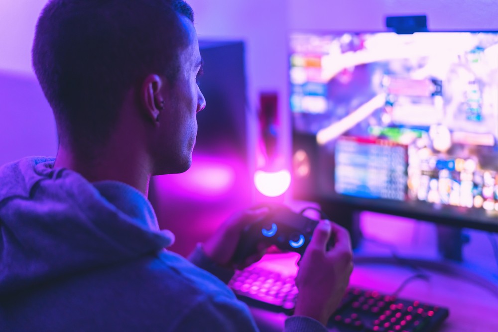 Young man sitting in front of computer gaming. The room is backit with a purple light.