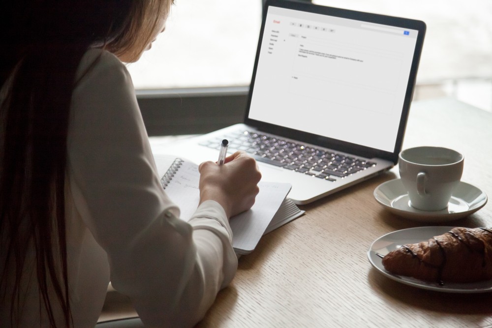 Woman sitting at a table with coffee, a croissant, and a laptop open in front of her to her email while she writes notes.