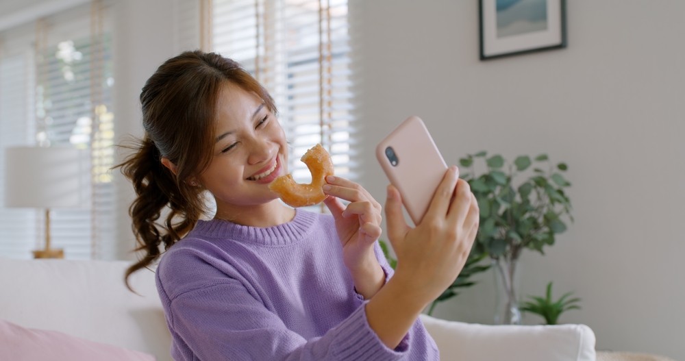 Asian girl in a purple sweater taking a selfie while holding up a half-eaten donut.