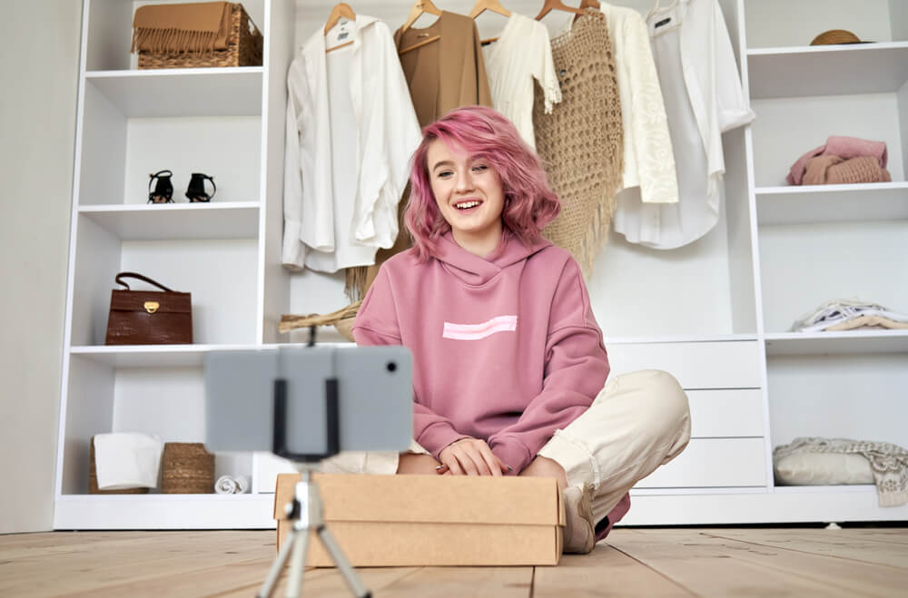 Pink-haired Gen Z girl sitting in front of a wardrobe unboxing while a phone records her on a tripod. 