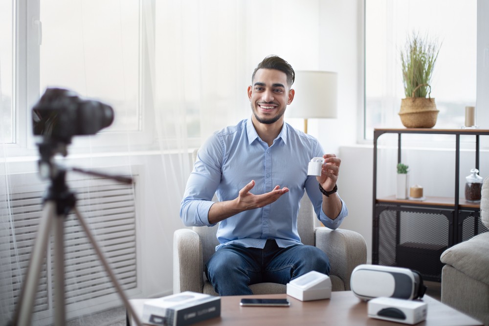 Hispanic yong man sitting in front of a tripod holding a set of earbuds. There are a variety of electronics sitting on the table in front of him.