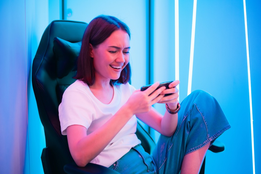 Young woman sitting in a gaming chair and messaging on her phone. Bright blue and white lights line her walls.