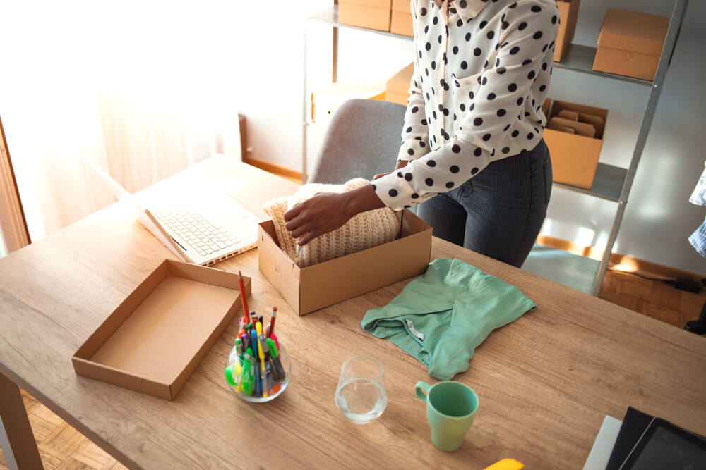 Woman wearing a polka dot blouse folding a sweater in a shipping box on her desk. Behind her is shipping supplies.