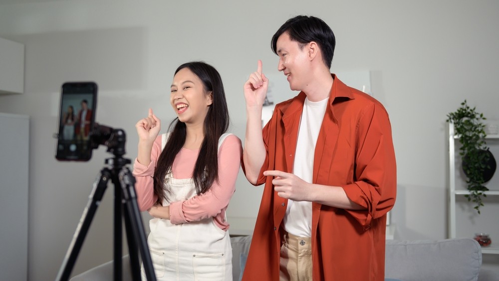 Two Asian people dancing in front of a cellphone on a tripod.