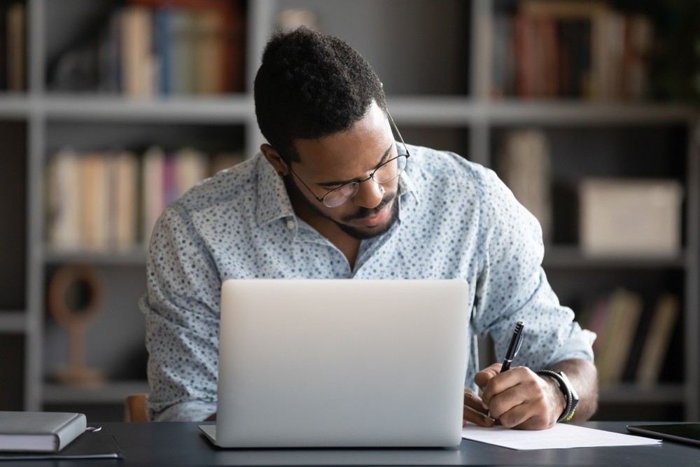 Black man wearing glasses and a dress shirt sitting in front of a lapop writing notes.