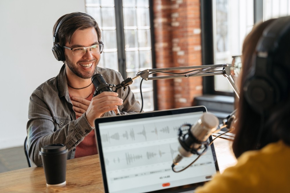 Man and woman sitting at a table with microphones and a laptop while recording a podcast.