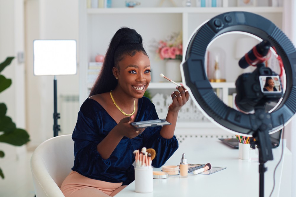 Young Black woman sitting in front of a ring light applying makeup.
