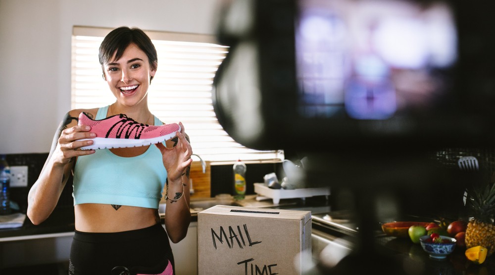 Woman in althetic apparel standing in front of a tripod recording herself holding up a shoe.w 