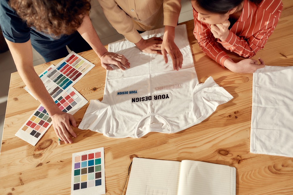 Three people leaning over a table designing a t-shirt with colore palettes and shirts sitting on the table.