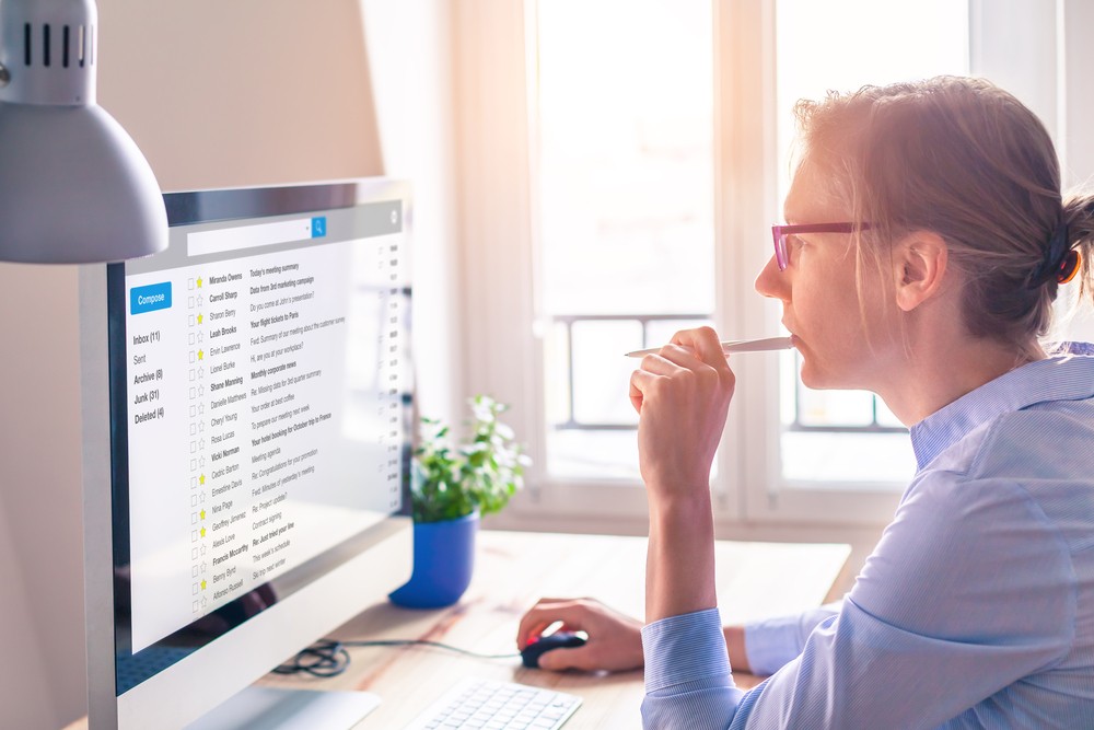 White woman sitting at her desk with a pen in her mouth checking email on a desktop computer.
