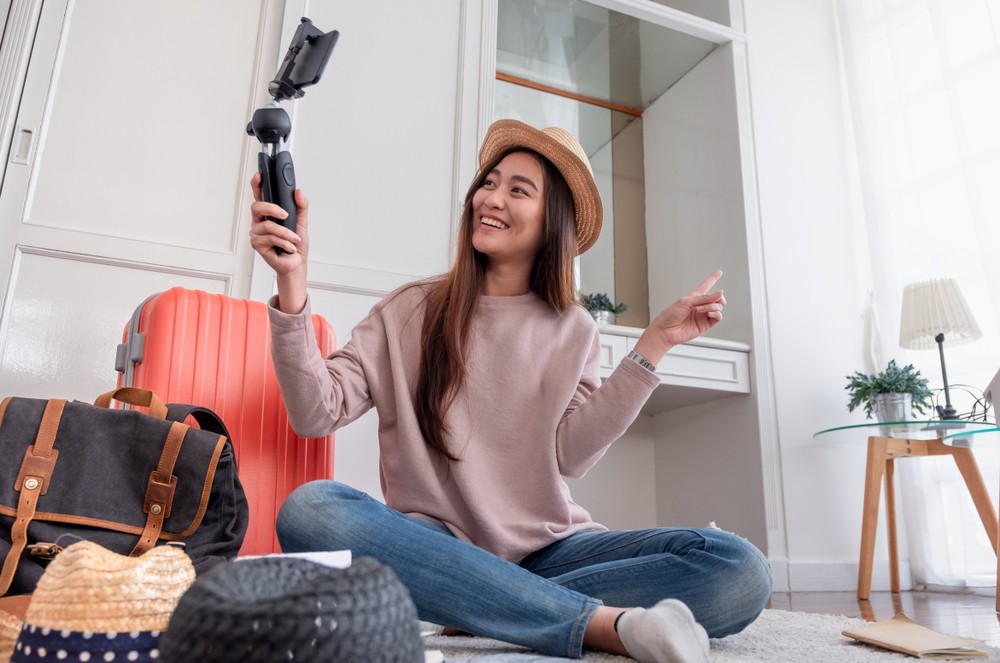 Asian woman sitting on her bedroom floor with luggage while filing herself on her phone.