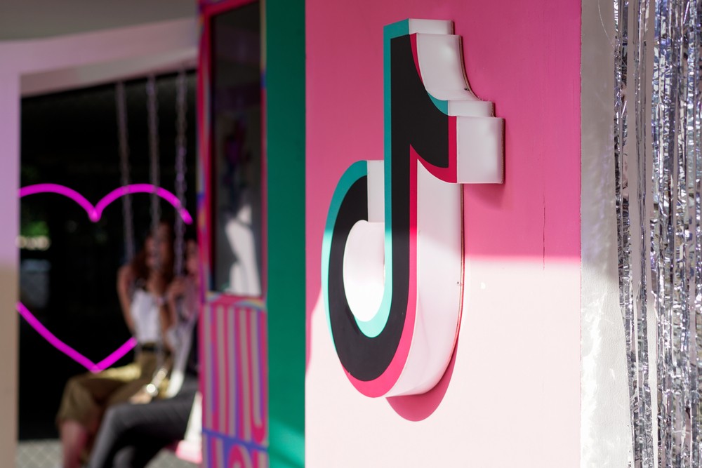 Closeup of the TikTok logo hanging on a pink wall with two women taking pictures on swings in the background.