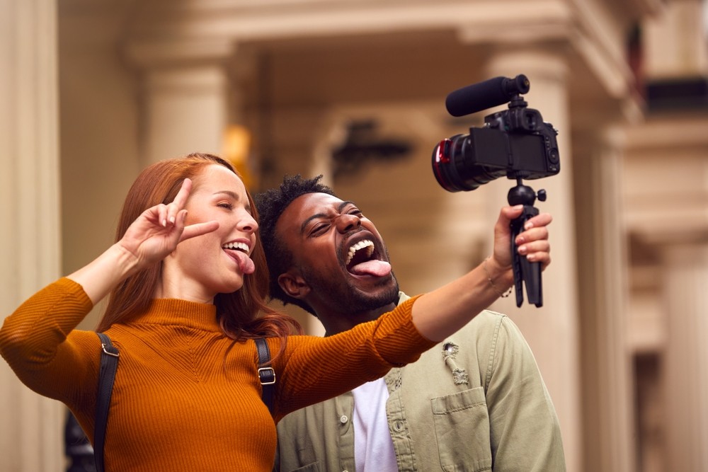 Young white red-haired woman holding up a peace sign with young black man sticking his tongue out and filming themselves with a tripod.