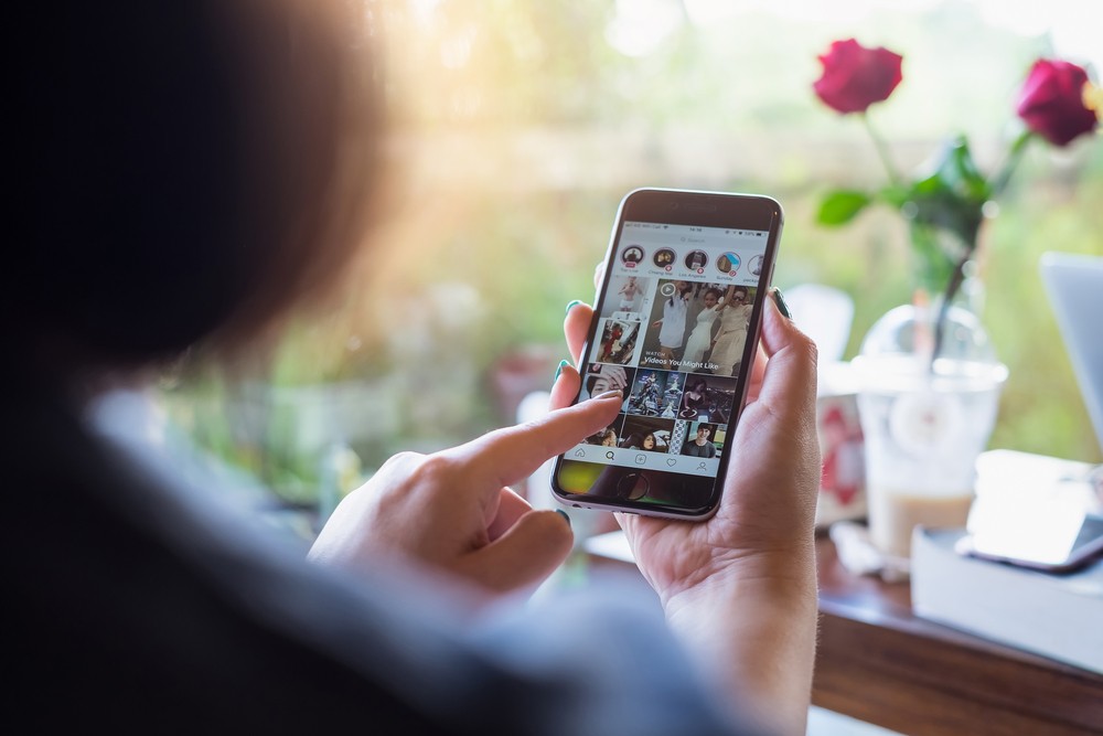 Closeup of woman holding her phone looking at Instagram explor epage with a coffee and flowers in the background.